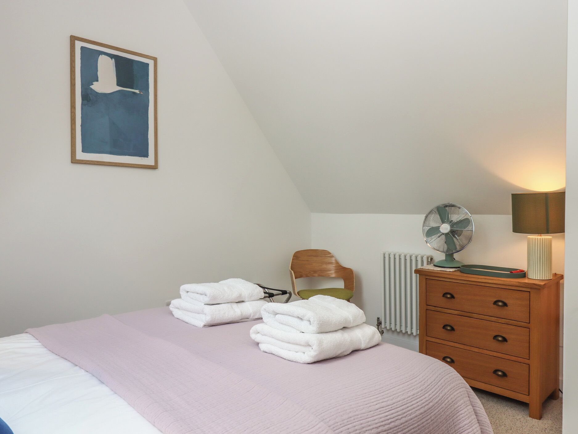 Ein Schlafzimmer mit einem Bett, einer hölzernen Kommode, einem Heizkörper, einem Ventilator und einem Bild an der Wand.