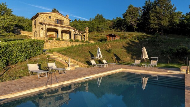 Une villa en pierre dotée d’une piscine, de chaises longues et d’une petite construction en bois, située sur le flanc d’une colline.