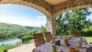 Un espace extérieur aménagé pour les repas, doté d’une voûte en pierre, et offrant une vue sur une piscine et des collines.