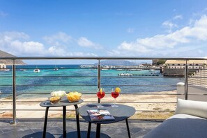 Un balcón con una mesa dispuesta para dos personas, con vistas a un puerto deportivo con barcos y un pueblo costero.