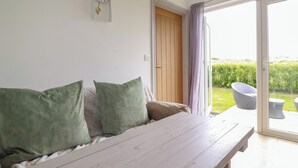 A room with a wooden bench, a green sofa, a wall clock, and a sliding glass door leading to an outdoor area with a garden and a wicker chair.