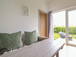A room with a wooden bench, a green sofa, a wall clock, and a sliding glass door leading to an outdoor area with a garden and a wicker chair. - The Tinystore (Bodmin)