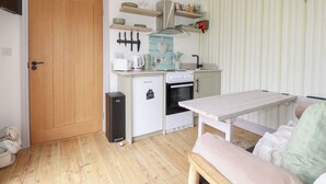 A compact kitchen with a refrigerator, stove, and microwave. There is a wooden door and a small table with a bench. - The Tinystore (Bodmin)