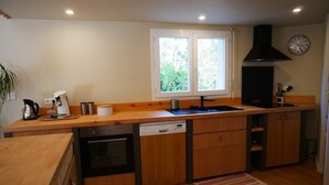 A compact kitchen with wooden cabinets, a dishwasher, a stove, and a window with a view of greenery. - Villa in the shade of the pines, 5 minutes from the beach. (Lacanau)