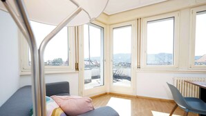 A modern room with a grey sofa, a wooden chair, and large windows offering a city view. - Spacious Zurich Apt, Huge Terrace & Views (Zürich)