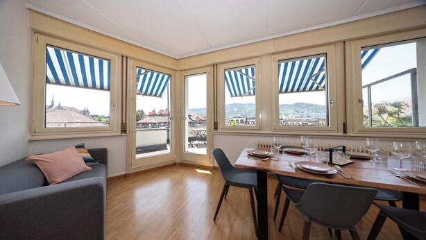 A dining area with a wooden table set for four, large windows with blue-striped awnings, and a gray sofa with a pink pillow. - Spacious Zurich Apt, Huge Terrace & Views (Zürich)
