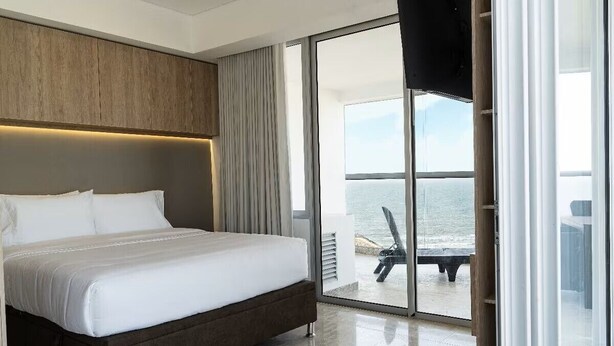 Una habitación de hotel moderna con una cama grande, cabecera de madera y puerta de cristal corredera que da a un balcón con vistas al mar.