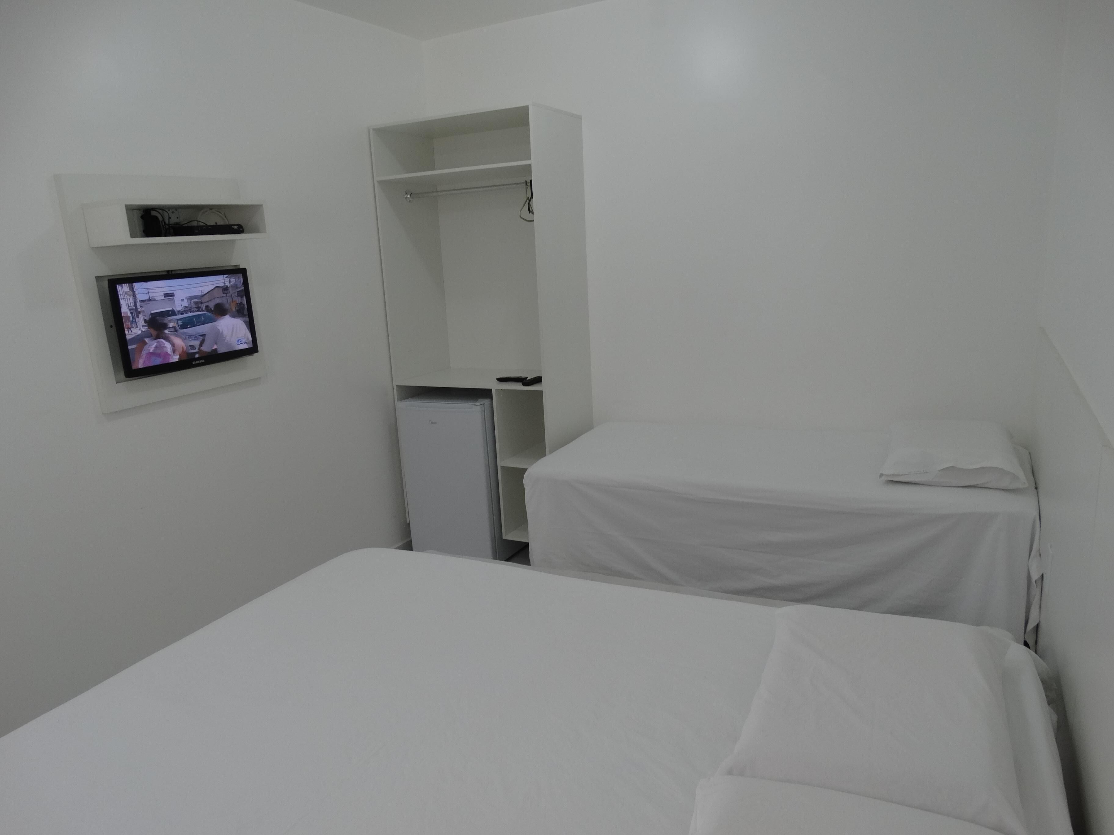 Une petite chambre d’hôtel blanche, composée d’une seule pièce, avec un lit, un téléviseur fixé au mur et une armoire.
