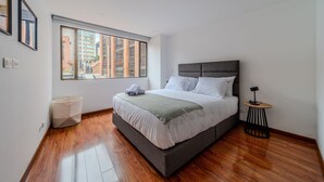 Quarto moderno com uma cama grande, janela com vista para a cidade e uma mesinha de cabeceira.