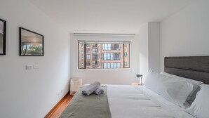 A modern bedroom with a large window, a bed with white linens, and a grey headboard.
