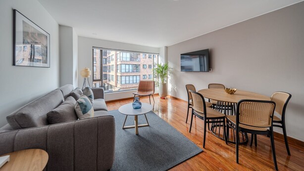A modern living room with a sofa, coffee table, dining table with chairs, a potted plant, and a TV mounted on the wall.