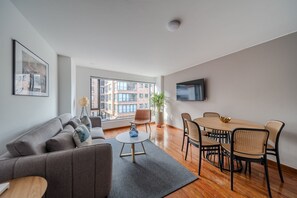 A modern living room with a sofa, coffee table, dining table with chairs, a potted plant, and a TV mounted on the wall.