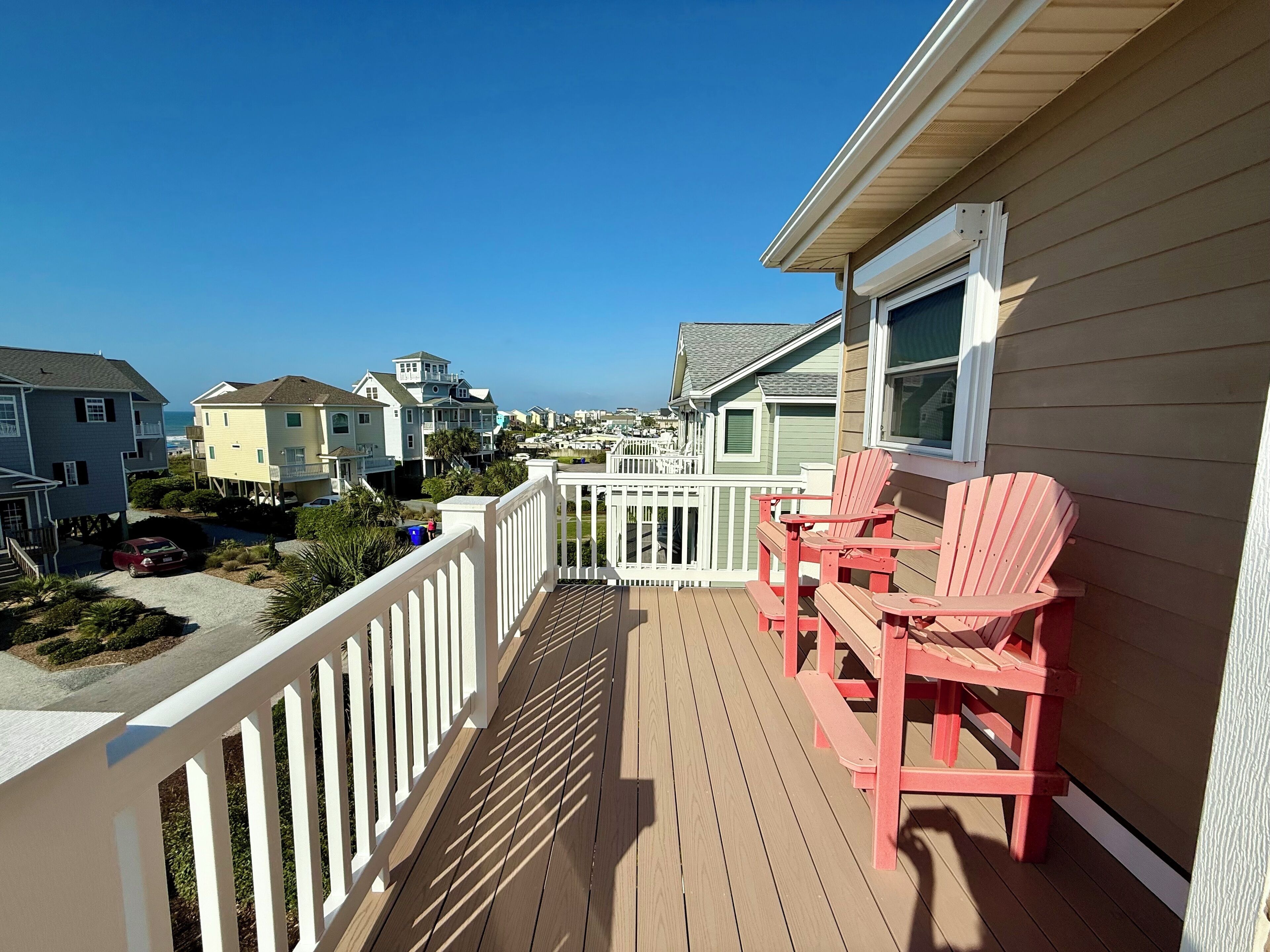 Une terrasse en bois avec une balustrade, deux chaises Adirondack rouges et une vue sur les maisons et l’océan.