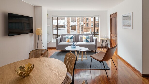 A modern living room with a sofa, coffee table, and chairs, a flat-screen TV mounted on the wall, and a large window offering a city view. - Bright 3BR @Commercial district with private terrace (Bogotá)
