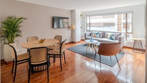 A modern living room with a wooden dining table, wicker chairs, a grey sofa with cushions, a TV mounted on the wall, and a large window providing natural light. - Bright 3BR @Commercial district with private terrace (Bogotá)