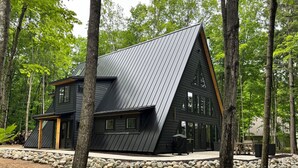 A modern, dark-colored cabin with a steeply pitched roof, surrounded by a stone retaining wall and tall trees. - Scenic 3-bedroom Aframe with WiFi and AC on Three Lakes Chain (Three Lakes)