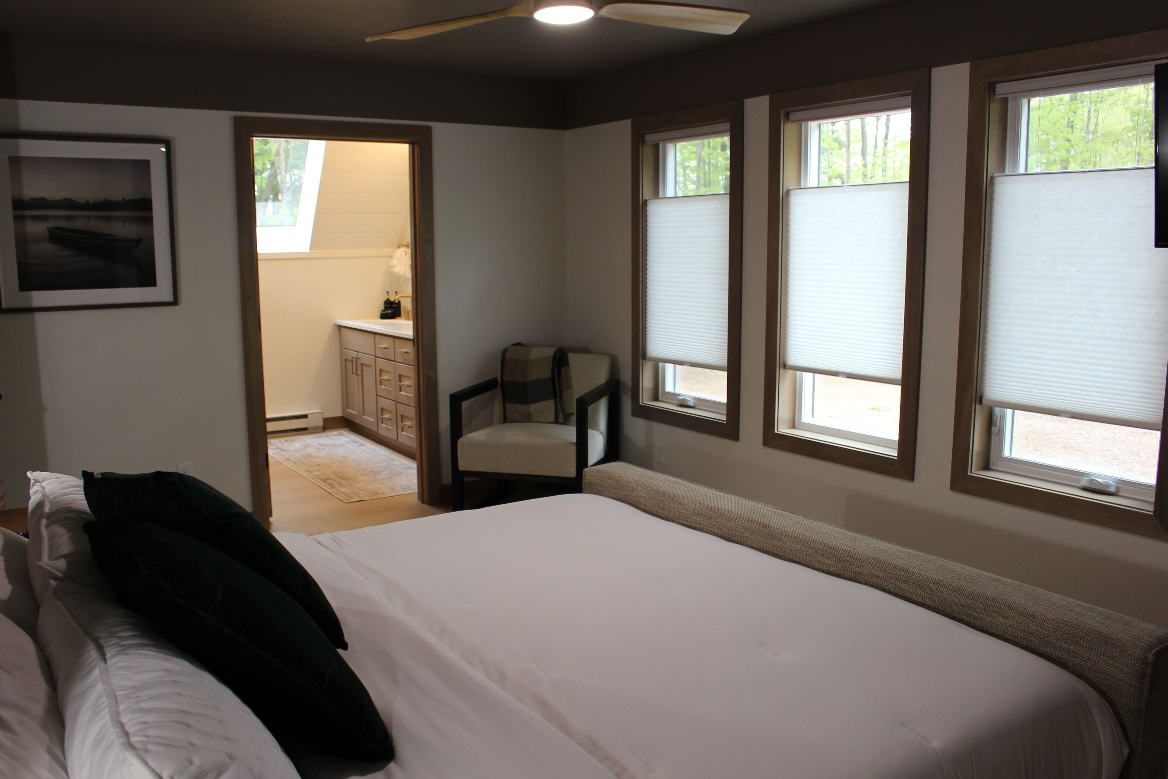 A bedroom with a large bed, a chair, and a desk. There are windows with blinds, a ceiling fan, and a framed picture on the wall.