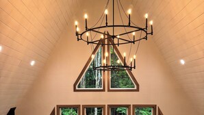 A room with a sloped ceiling, a chandelier, and large windows offering a view of a wooded area. - Scenic 3-bedroom Aframe with WiFi and AC on Three Lakes Chain (Three Lakes)