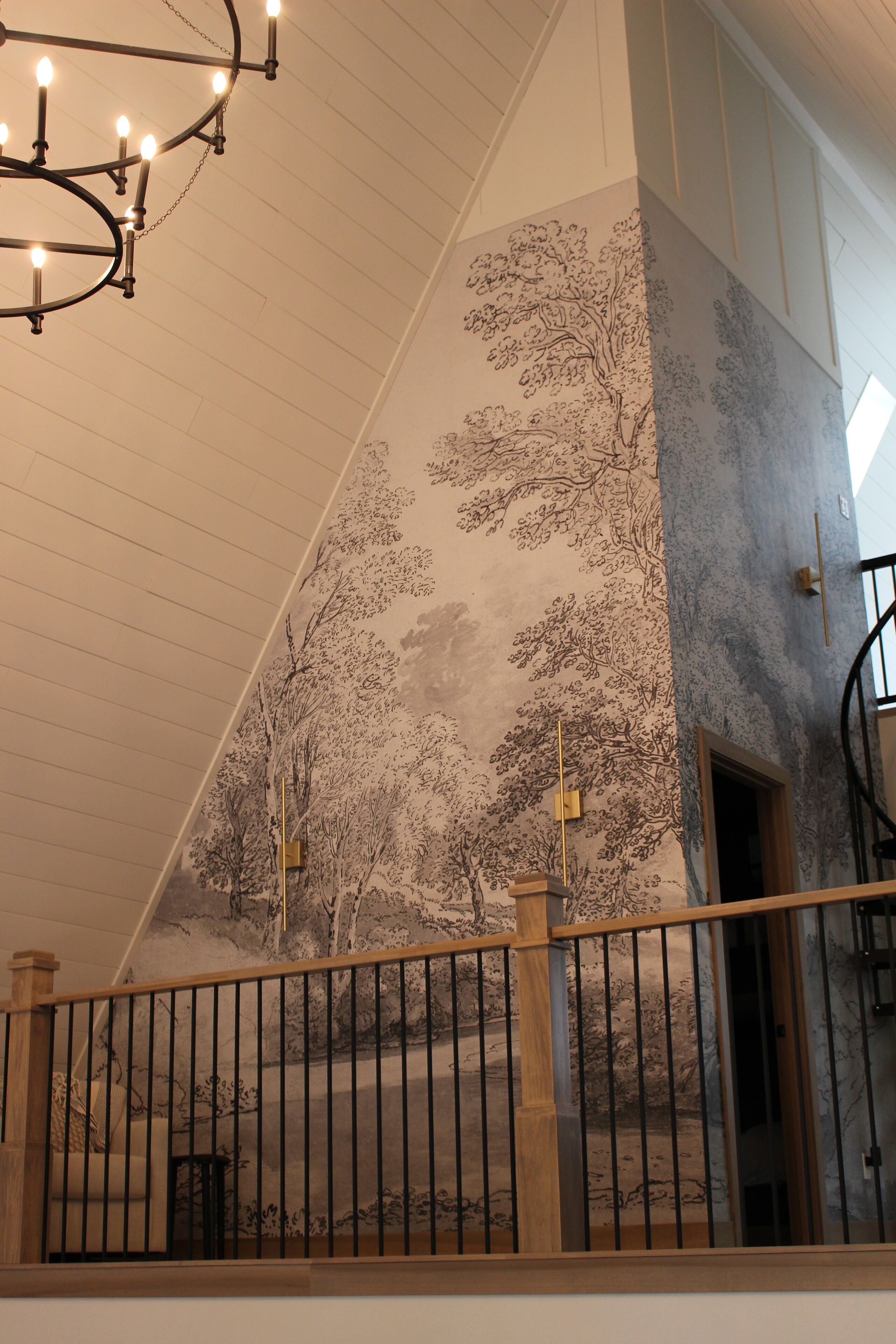 A staircase with a wooden handrail and a black metal railing, leading to a room with a high ceiling and a chandelier.