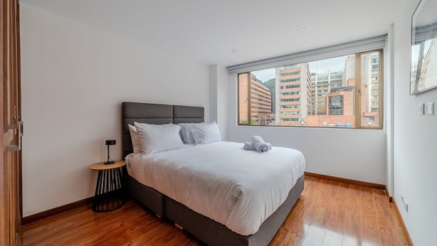 Un dormitorio moderno con una cama grande, una mesita auxiliar pequeña y una ventana con vistas a la ciudad.