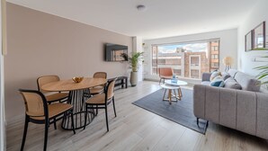 Un salon moderne avec un canapé gris, une table à manger en bois avec des chaises, une grande fenêtre donnant sur des immeubles et un téléviseur à écran plat fixé au mur.