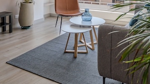 Un salon moderne avec une grande fenêtre, une plante en pot, un fauteuil en cuir marron, une table ronde avec un vase bleu et un tapis gris.