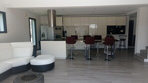 Una sala moderna con un sofá, un reposapiés y una mesa de centro. Se observan cuatro taburetes de bar y una mesa de comedor al fondo.