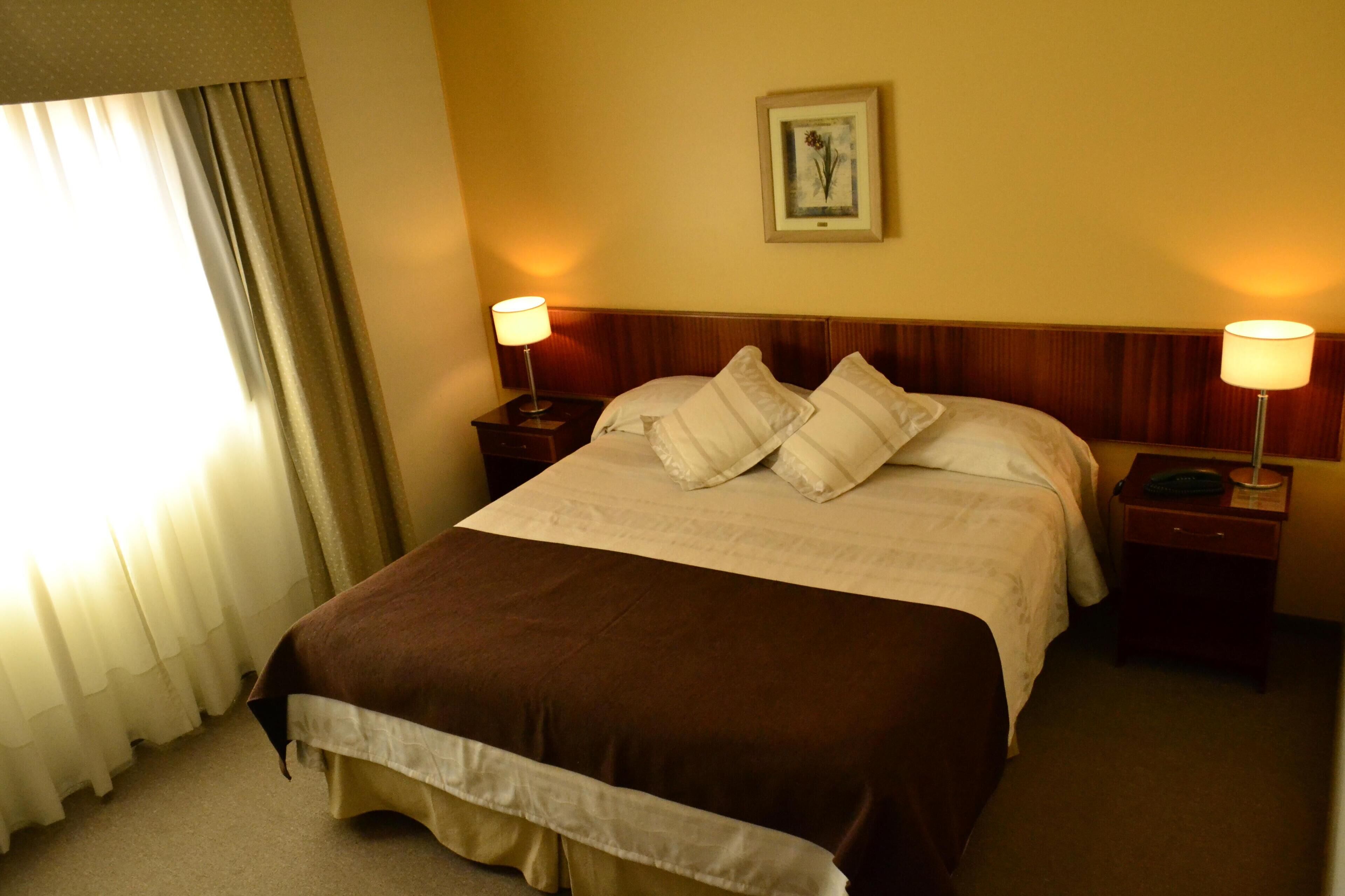 Een hotelkamer met een groot bed, twee nachtkastjes, een raam met gordijnen en een lijst met een afbeelding aan de muur.
