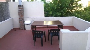 A small outdoor patio with a wooden table and two chairs, a trash bin, and a brick chimney. - Hostel Casa la Red (Mar del Plata)