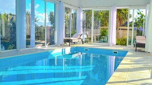 A swimming pool with clear blue water, surrounded by large windows and a covered patio area with lounge chairs.