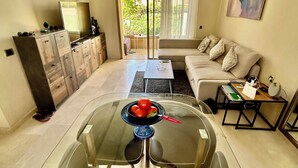 Een moderne woonkamer met een glazen eettafel, een bank, een televisie en een balkon met gordijnen.