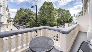 Un balcone con tavolo e sedie, affacciato su una strada con auto parcheggiate e alberi.