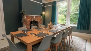 A dining room with a long wooden table set for six, surrounded by grey chairs. A brick fireplace is visible in the background, and the room has a large window with teal curtains. - Montclair (Buxton)
