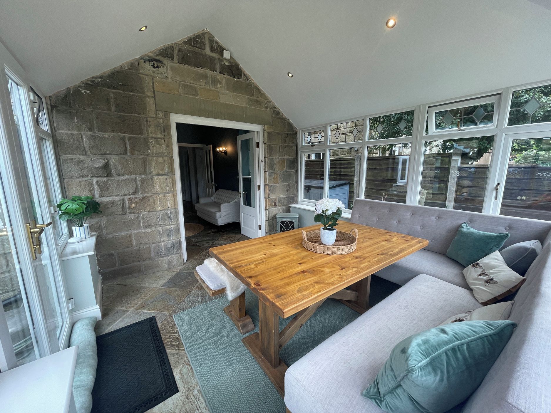 A cozy room with a stone wall, a wooden table, a grey sofa, and a window with stained glass.