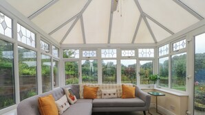 A conservatory with a grey sectional sofa, a small table, and a view of a garden through large windows. - Montclair (Buxton)