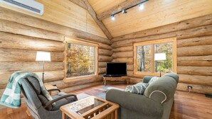 A log cabin interior with a sofa, chair, TV, and large windows. - Stagecoach Inn Log Cabin 10 Acres Wooded Setting (Beulah)