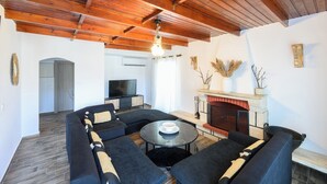A living room with a fireplace, dark sofas, a TV, and wooden ceiling beams. - Azure Horizon Skyros (Aspous)