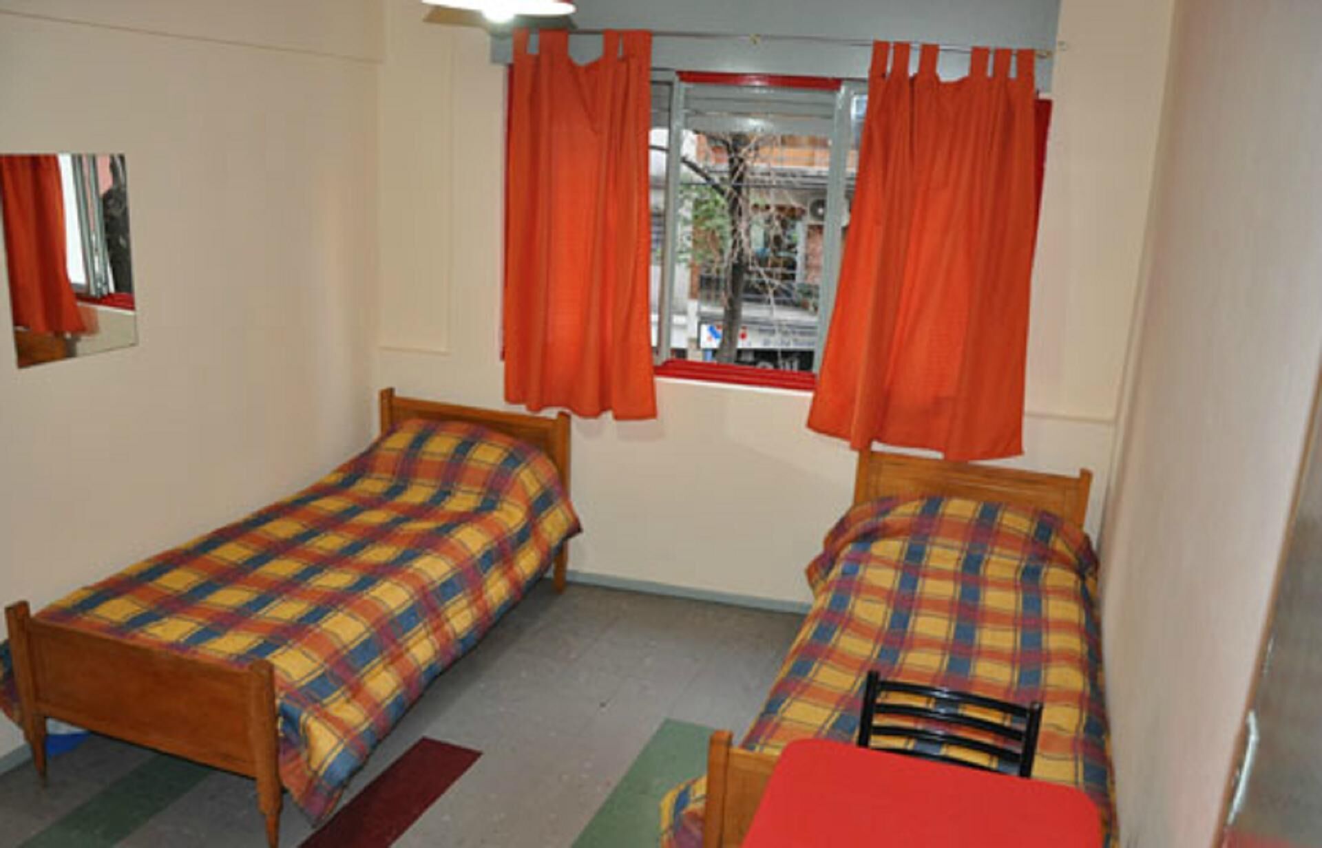 Una habitación con dos camas de madera, cada una con cubrecama a cuadros, y una ventana con cortinas naranjas.