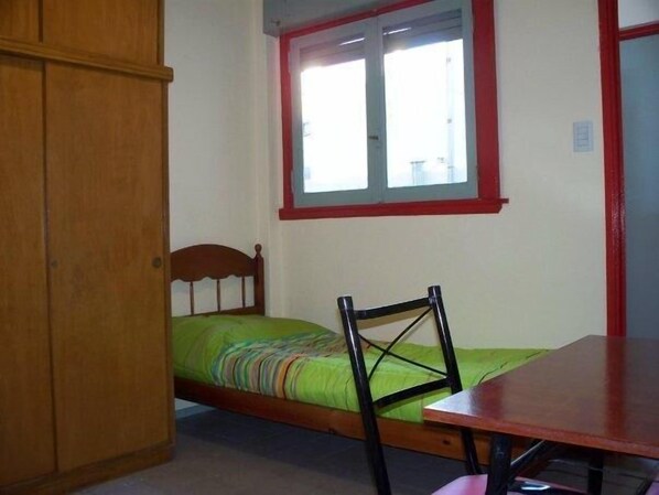 A room with a bed, a wooden wardrobe, a table, and a window with red trim.