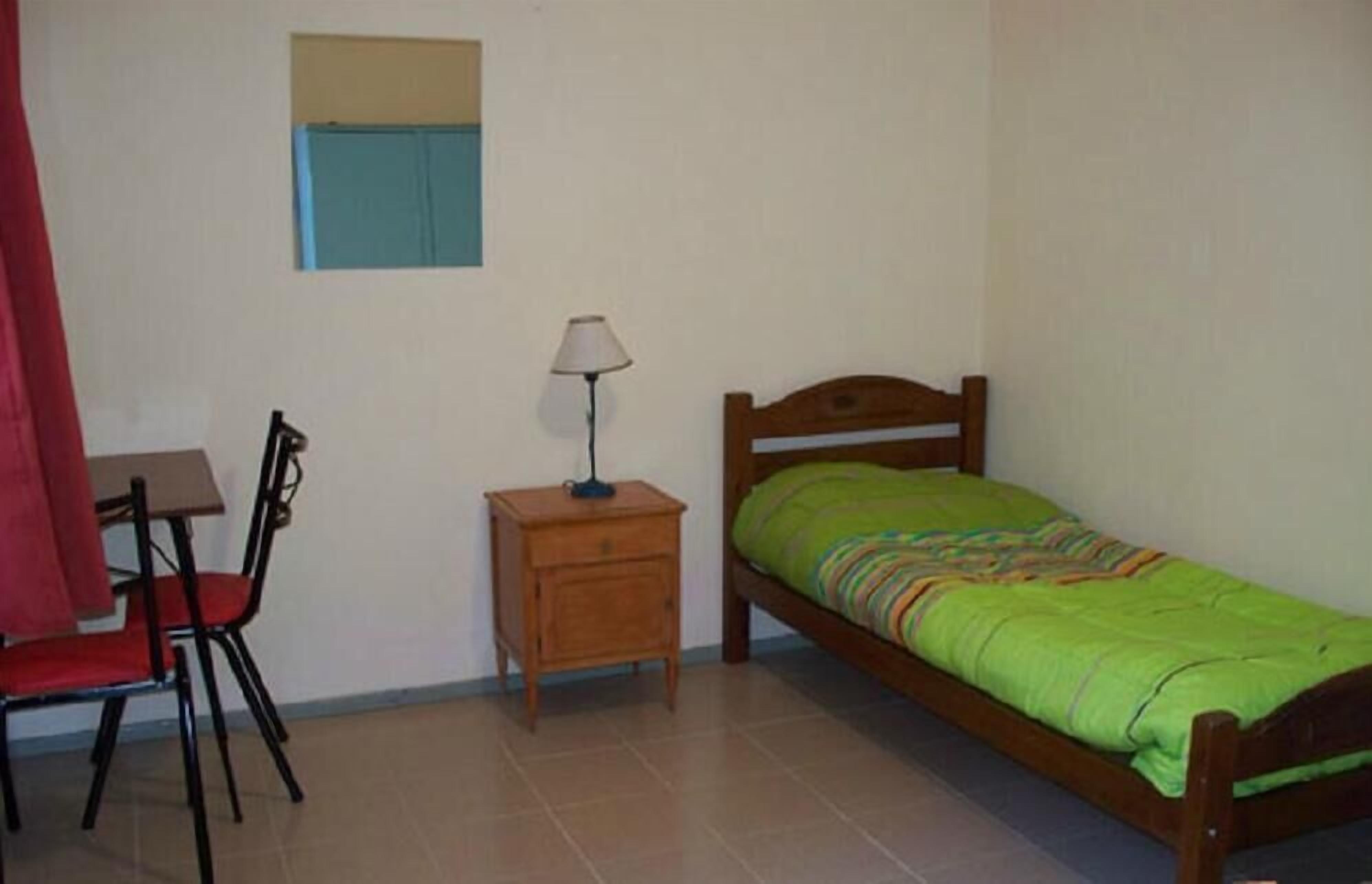 A single bed with a green blanket, a small bedside table with a lamp, a chair, and a desk with a red chair.