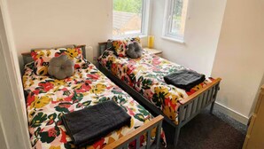 A small, two-bed room with floral bedding, a grey pillow, and a black blanket on each bed. There are two windows with black blinds, and a radiator on the right side. - Modern 3 bed home next to railway station (North Ferriby)