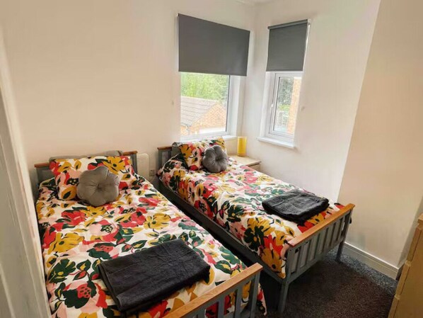 A small, two-bed room with floral bedding, a grey pillow, and a black blanket on each bed. There are two windows with black blinds, and a radiator on the right side. - Modern 3 bed home next to railway station (North Ferriby)
