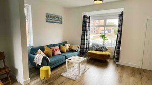A living room with a blue sofa, a yellow ottoman, a white coffee table, and a window with curtains. - Modern 3 bed home next to railway station (North Ferriby)