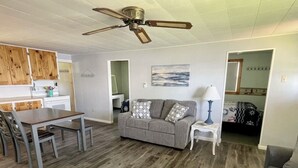 Un salon avec un canapé gris, une table à manger avec des chaises, un ventilateur de plafond et un coin cuisine doté de meubles en bois.
