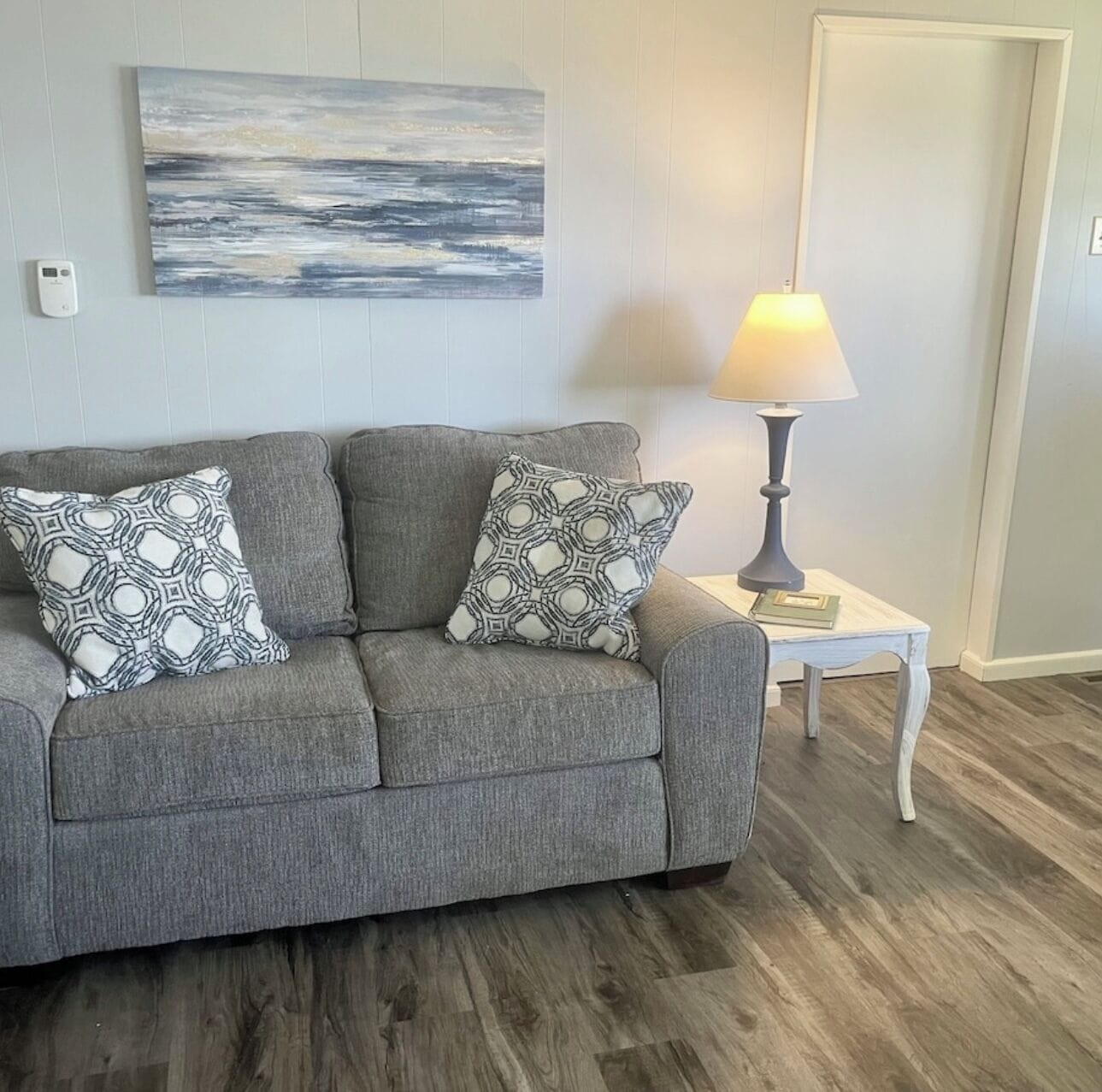 A grey sofa with patterned cushions, a wall-mounted abstract painting, a table lamp, and a wooden floor.