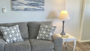 Un canapé gris agrémenté de coussins à motifs, un tableau abstrait accroché au mur, une lampe de table et un plancher en bois.