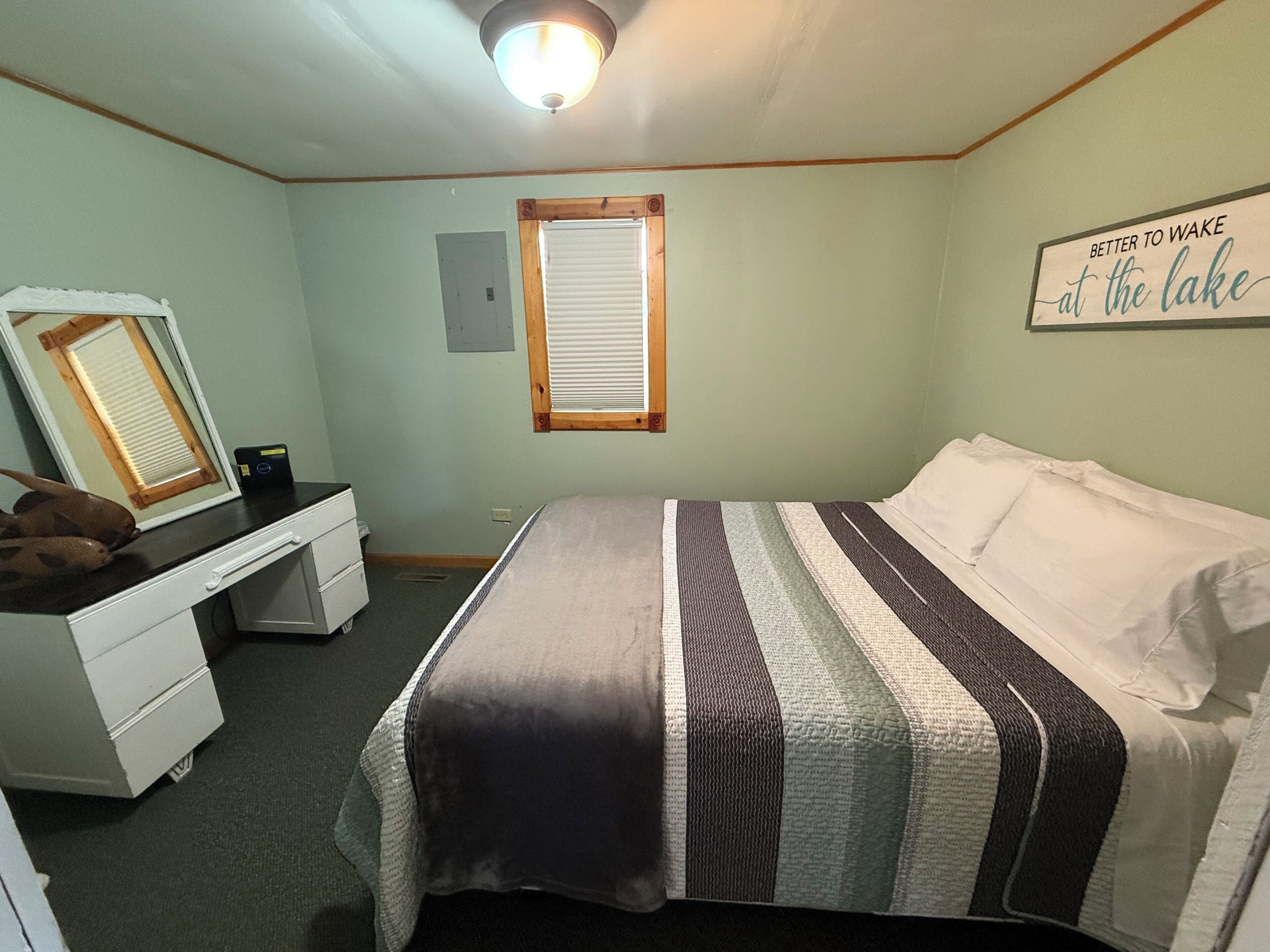Une chambre à coucher comprenant un lit, une commode, un miroir, une fenêtre avec des stores et un tableau mural.
