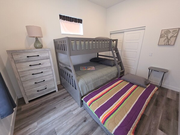 A bedroom with a bunk bed, a dresser, a lamp, a window with a valance, a door, and a small table.