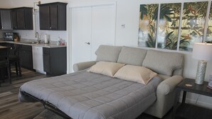A bedroom with a bed, a nightstand, a lamp, a chair, a desk, and a kitchen area with cabinets and appliances.