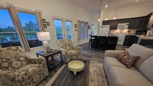 A modern living room with patterned furniture, a coffee table, and a sofa. There is a kitchen area with dark cabinets and a dining space with chairs.